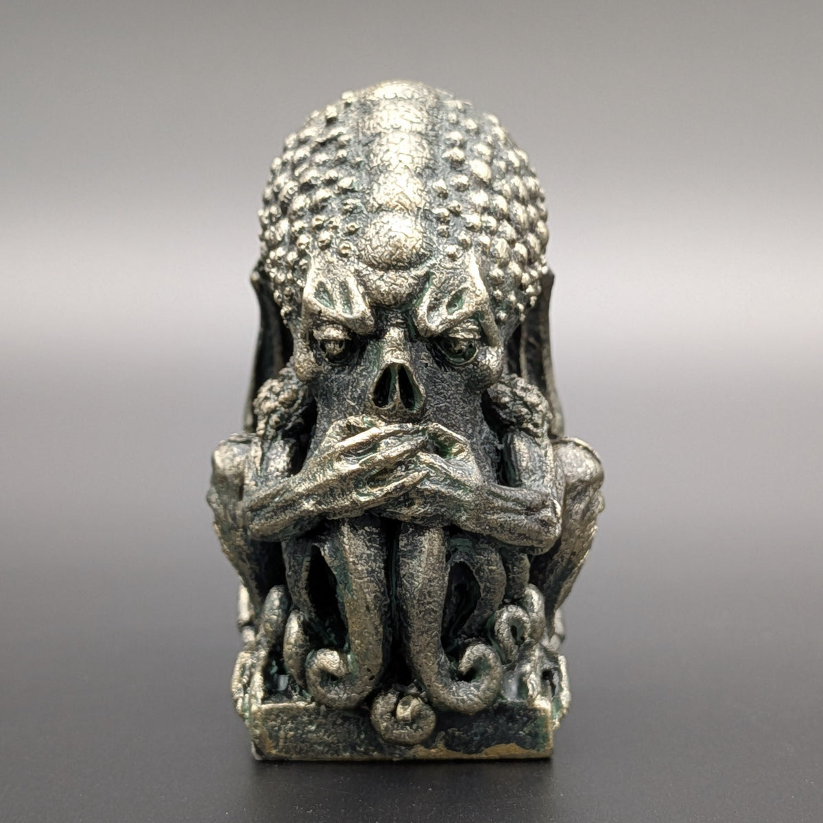 A polyresin Cthulhu figurine with a weathered stone patina & textured skin, depicting "Speak no evil" by covering his mouth, displayed on a dark grey background.