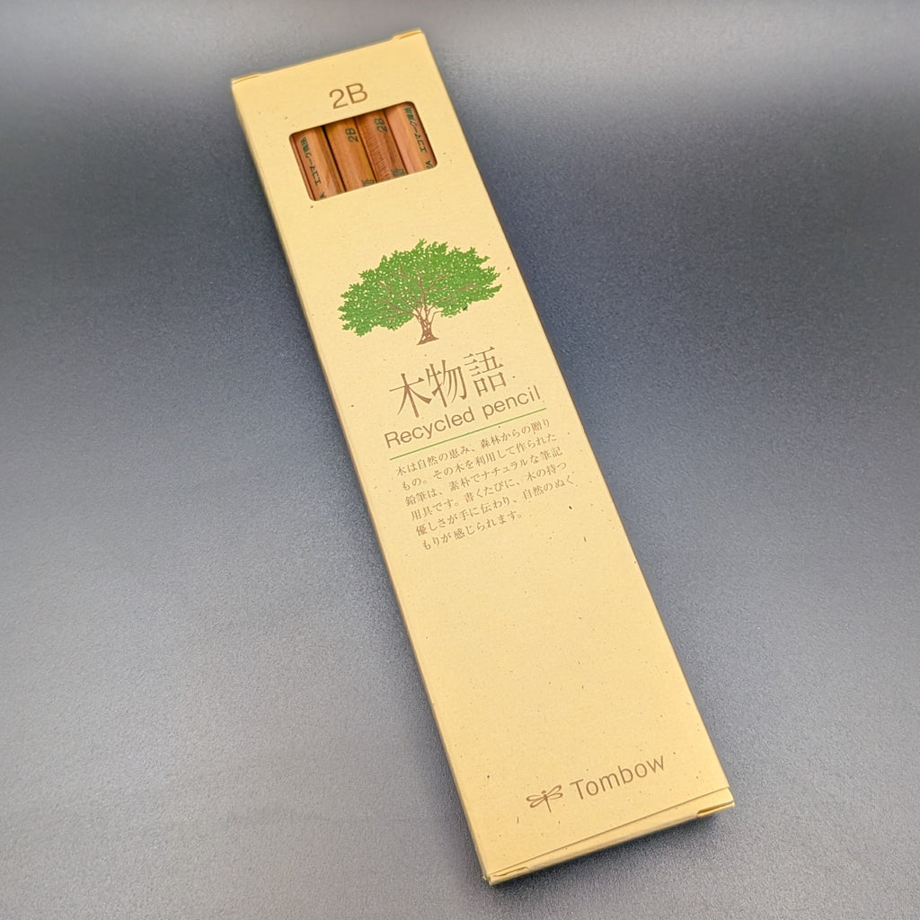 A tall, narrow beige cardboard box for Tombow 2B Recycled pencil, featuring a green tree illustration and Japanese text, set against a dark grey background. The box is leaning to the left.