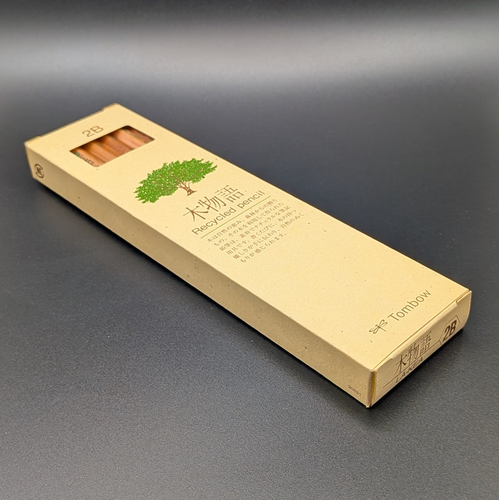 A tall, narrow beige cardboard box for Tombow 2B Recycled pencil, featuring a green tree illustration and Japanese text, laying on its back, set against a dark grey background.