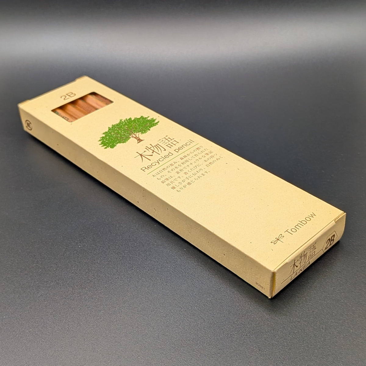 A tall, narrow beige cardboard box for Tombow 2B Recycled pencil, featuring a green tree illustration and Japanese text, laying on its back, set against a dark grey background.