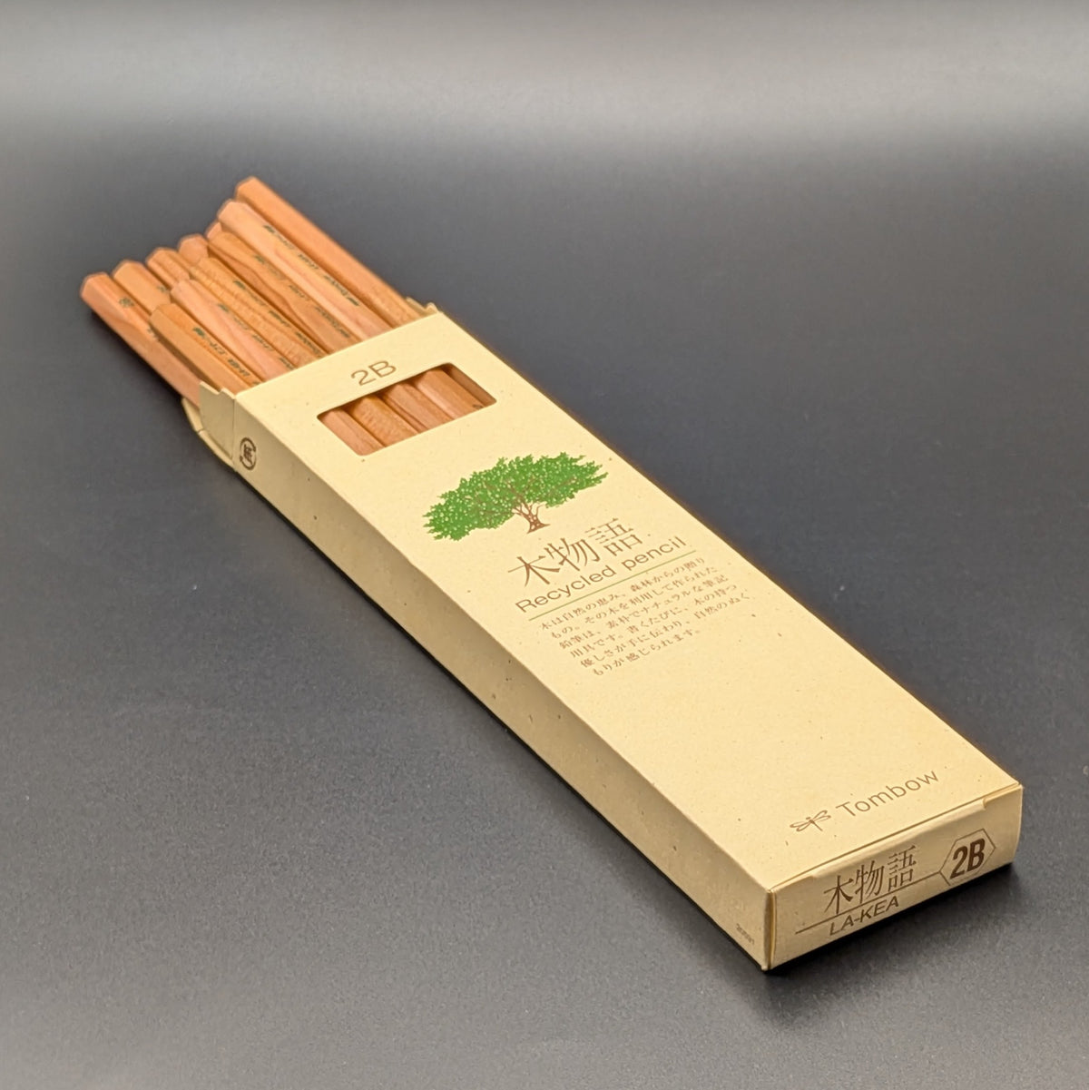 A tall, narrow beige cardboard box for Tombow 2B Recycled pencil, featuring a green tree illustration and Japanese text, half open revealing the pencils in neat rows, set against a dark grey background.