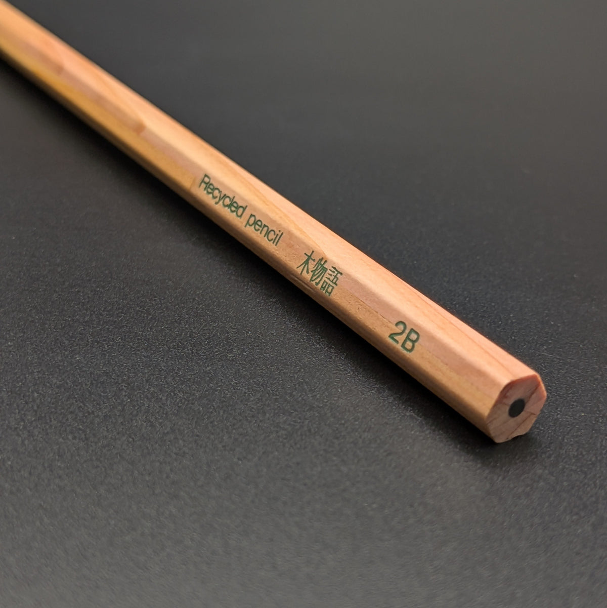 A close-up of a wooden pencil with green text reading 'Recycled pencil' and '2B' on a dark textured background.