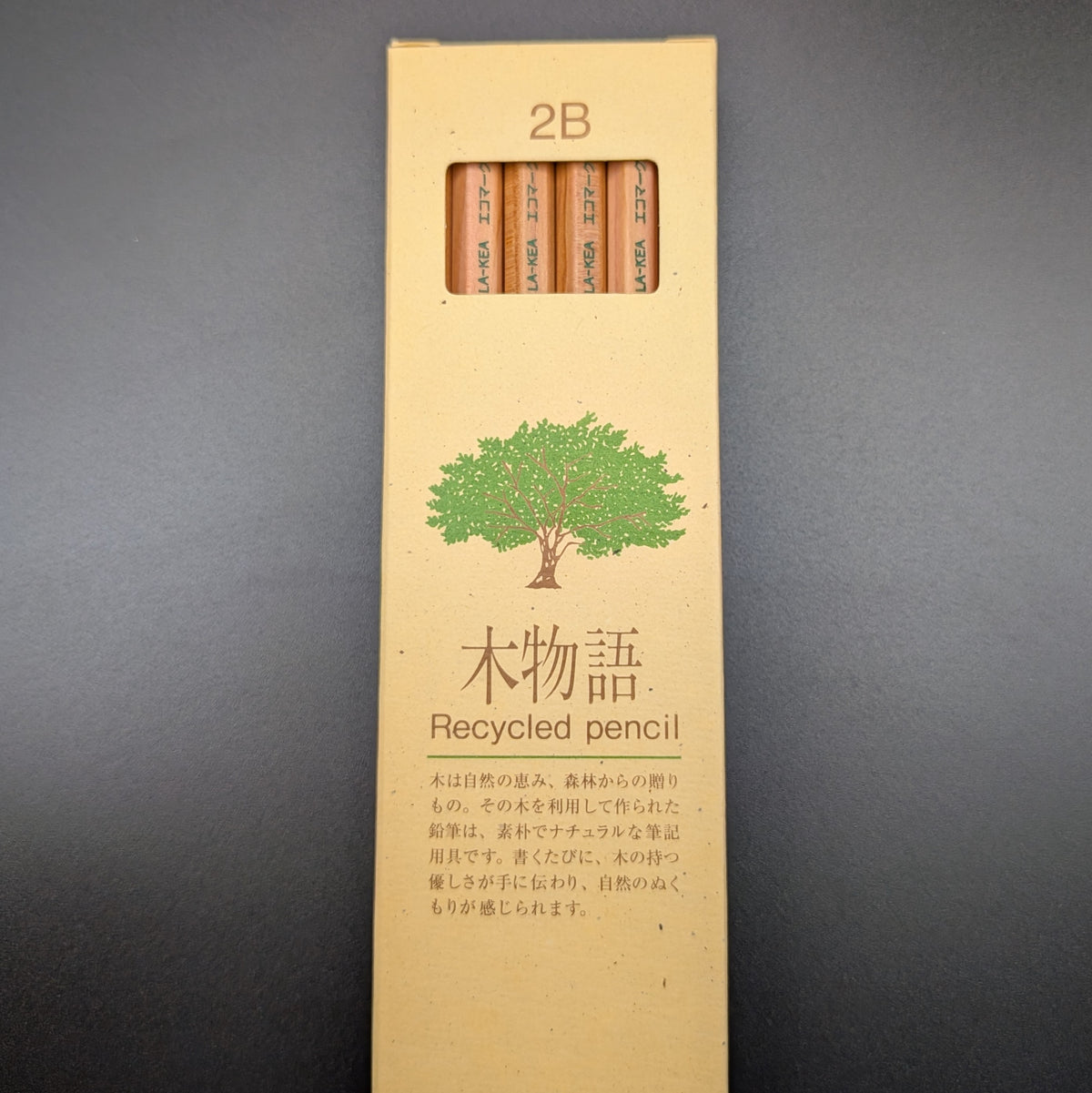 A tall, narrow beige cardboard box for Tombow 2B Recycled pencil, featuring a green tree illustration and Japanese text, set against a dark grey background. Standing up and facing forward.