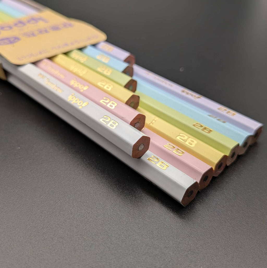 A set of pastel-colored wooden pencils arranged diagonally on a dark gray surface, with a tan paper tag partially visible at the top left.