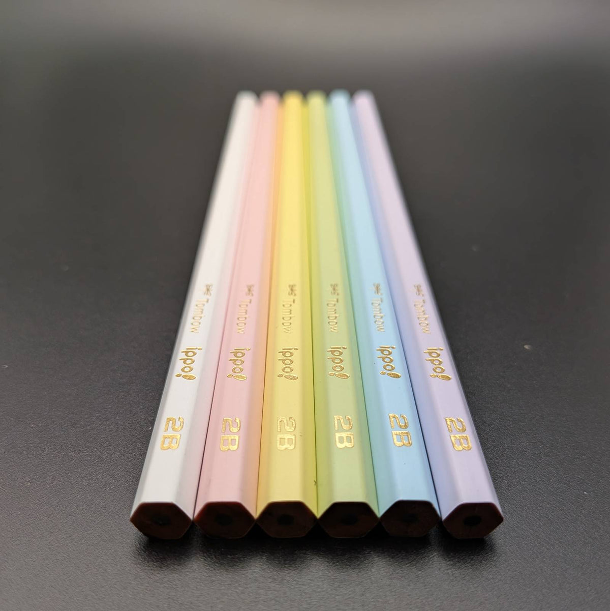 Pastel-colored pencils arranged on a dark gray surface. The pencils are white, pink, yellow, green, blue, and purple, each printed with the brand name 'ippo!' and the model '2B'.