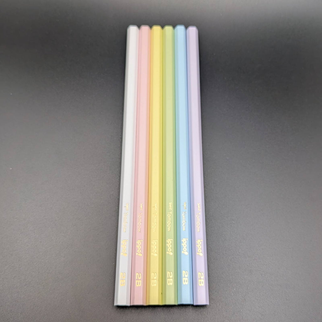 Six pastel-colored pencils standing upright in a vertical row on a dark gray background. The pencil colors are white, pink, yellow, green, blue, and purple, with gold text near the tips.