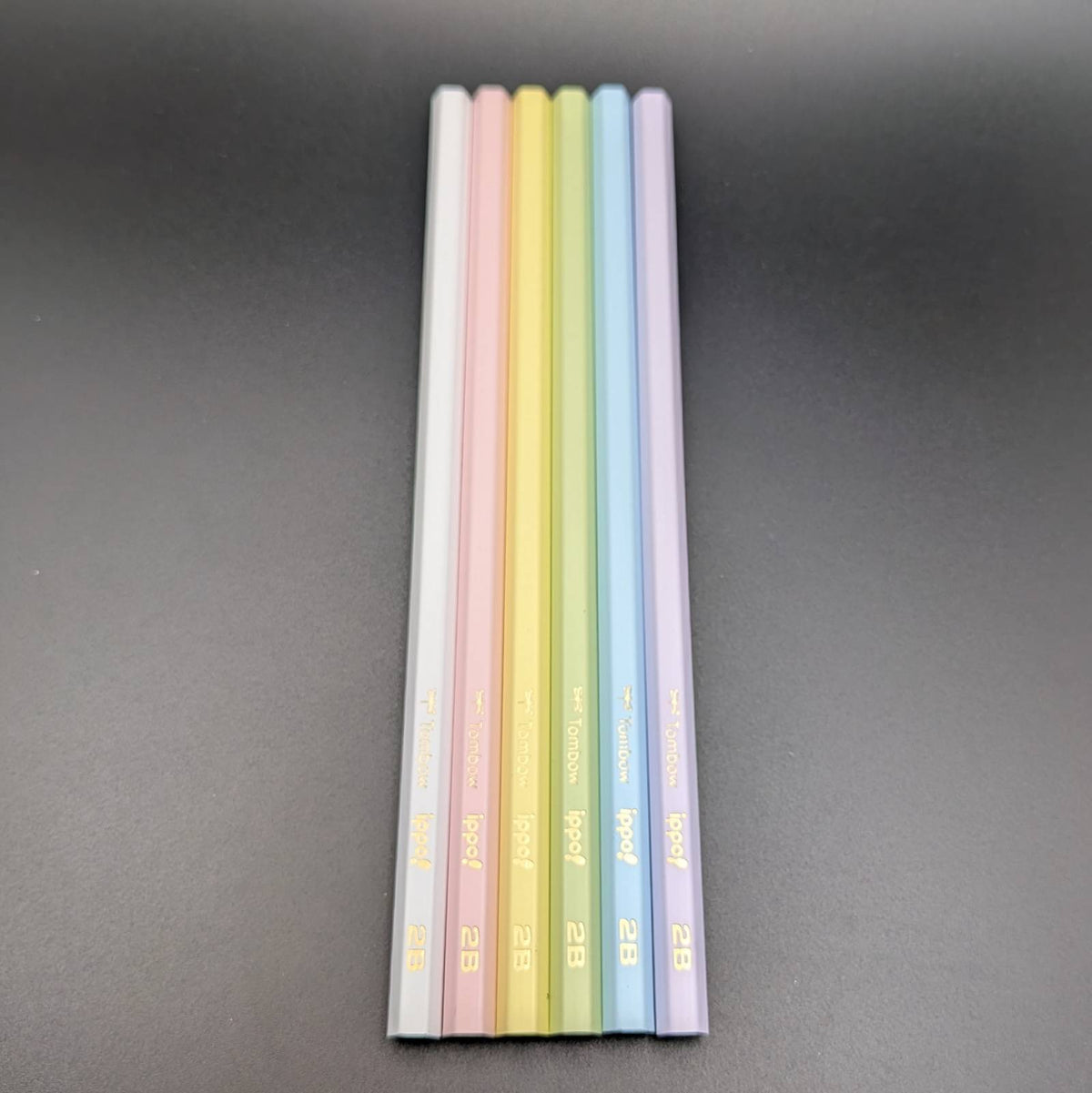 Six pastel-colored pencils standing upright in a vertical row on a dark gray background. The pencil colors are white, pink, yellow, green, blue, and purple, with gold text near the tips.