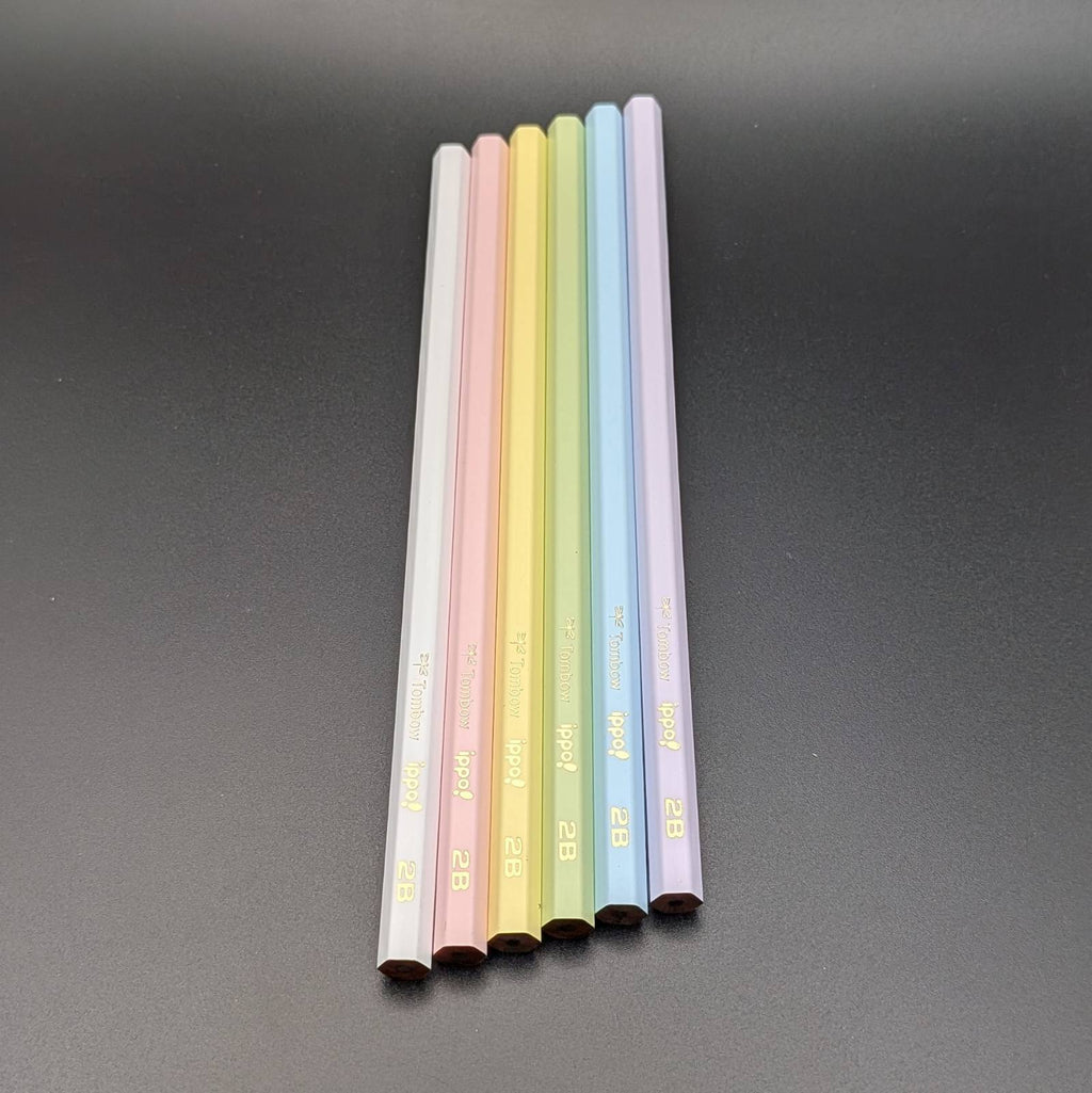 Six pastel-colored pencils standing upright in a row on a dark gray surface. The pencil colors are white, light pink, yellow, light green, light blue, and light purple, each with gold text near the bottom.