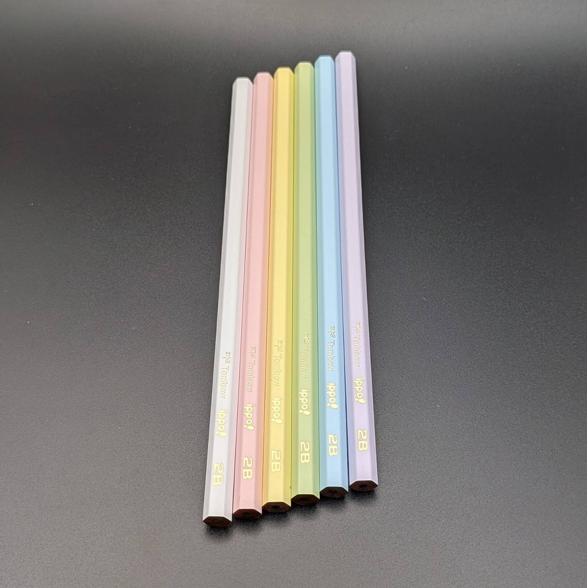 Six pastel-colored pencils standing upright in a row on a dark gray surface. The pencil colors are white, light pink, yellow, light green, light blue, and light purple, each with gold text near the bottom.