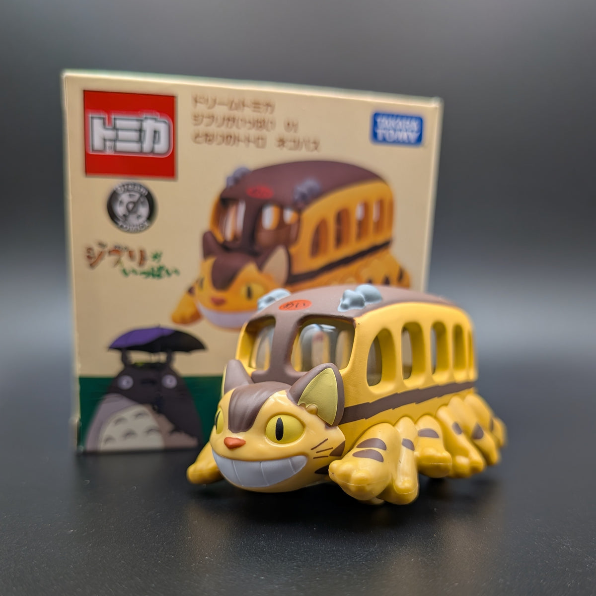 A small yellow and brown Catbus toy from Totoro's stands in front of its original Japanese packaging box on a dark gray surface.