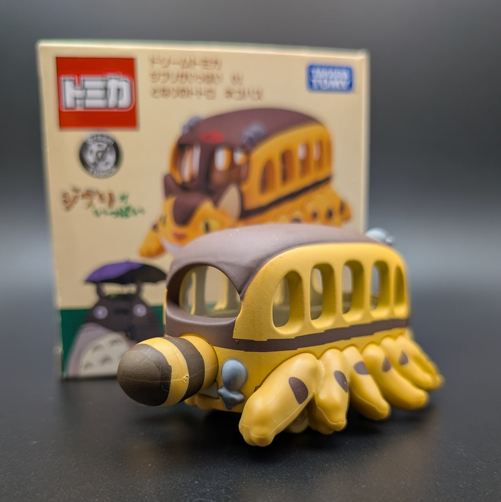 A small yellow and brown Cat Bus figure viewed from behind with multiple legs, shown in front of its product box. The box features the Takara Tomy logo and an illustration of the model.