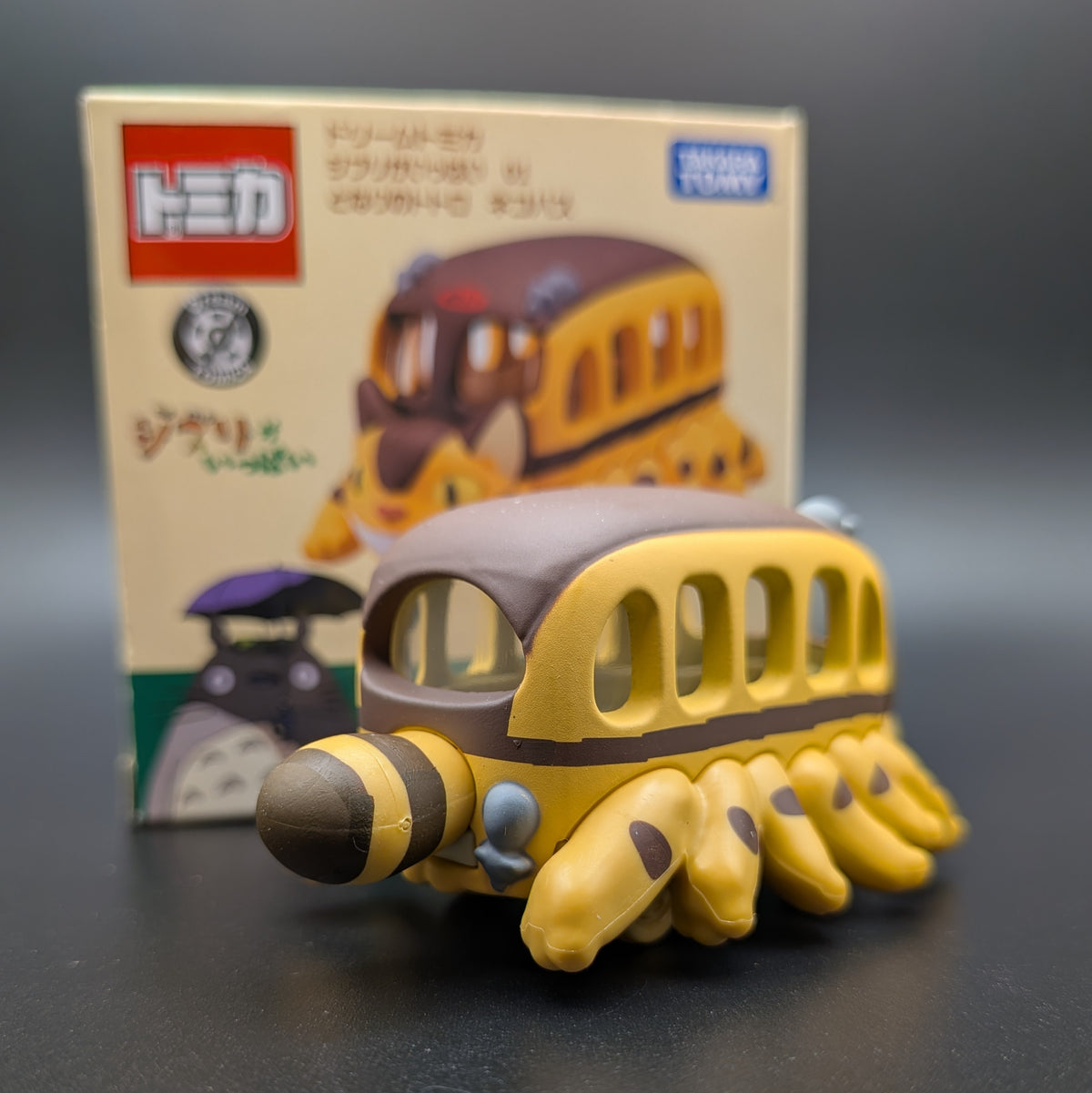 A small yellow and brown Cat Bus figure viewed from behind with multiple legs, shown in front of its product box. The box features the Takara Tomy logo and an illustration of the model.