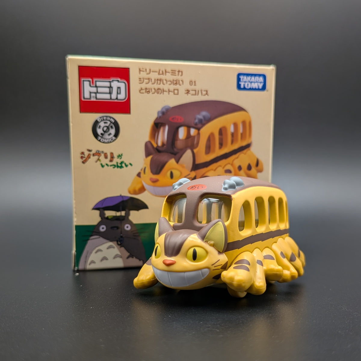 A small yellow and brown Totoro-themed toy bus figure placed in front of its branded retail box on a dark gray surface, with the box showing Japanese text and Tomy branding.