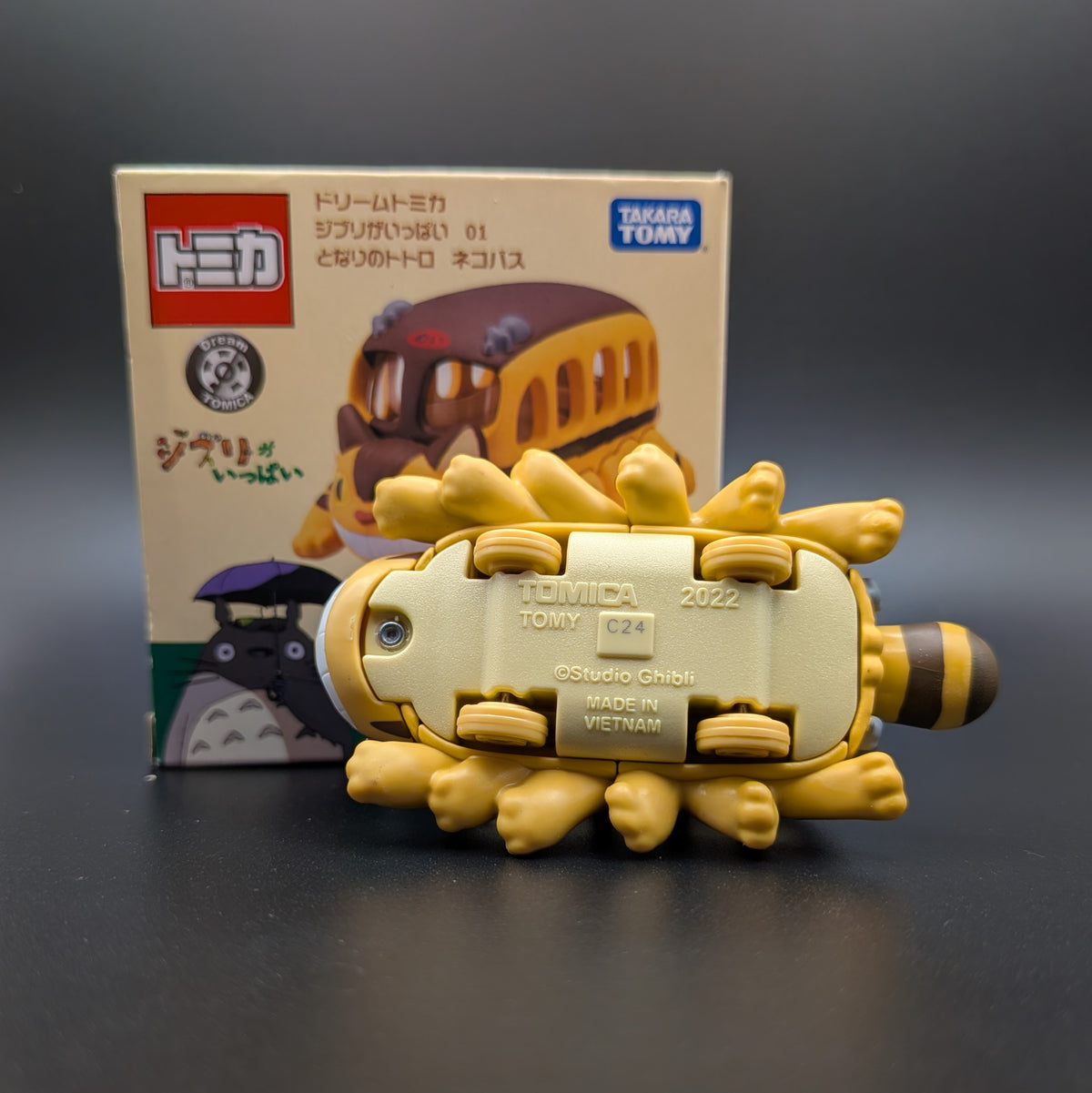 Underside view of the Takara Tomy Cat Bus from My Neighbour Totoro, displayed in front of its original retail packaging on a dark gray surface.