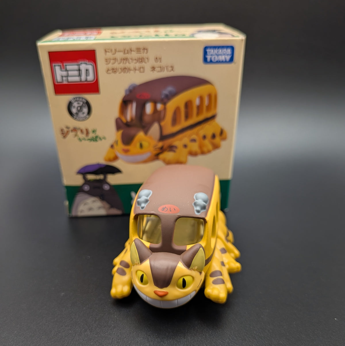 Tomica collectible toy bus shaped like Totoro's cat bus, placed in front of its original Japanese Tomy box on a dark gray background.