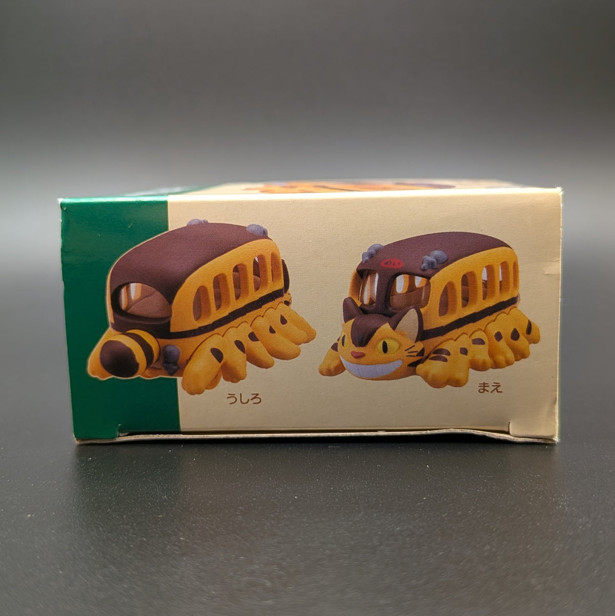 A small toy bus figurine shown on its printed retail box. The box features illustrations of the toy from two angles, with a green side panel and a cream front panel.