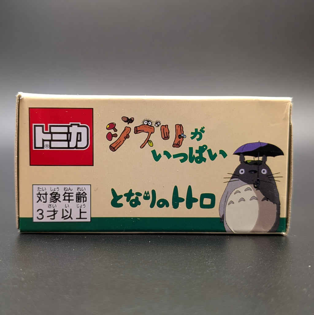 A boxed product on a dark gray surface with a gray gradient background. The box is beige with a red Tomica logo, Japanese text, and an illustration of Totoro holding a purple umbrella.