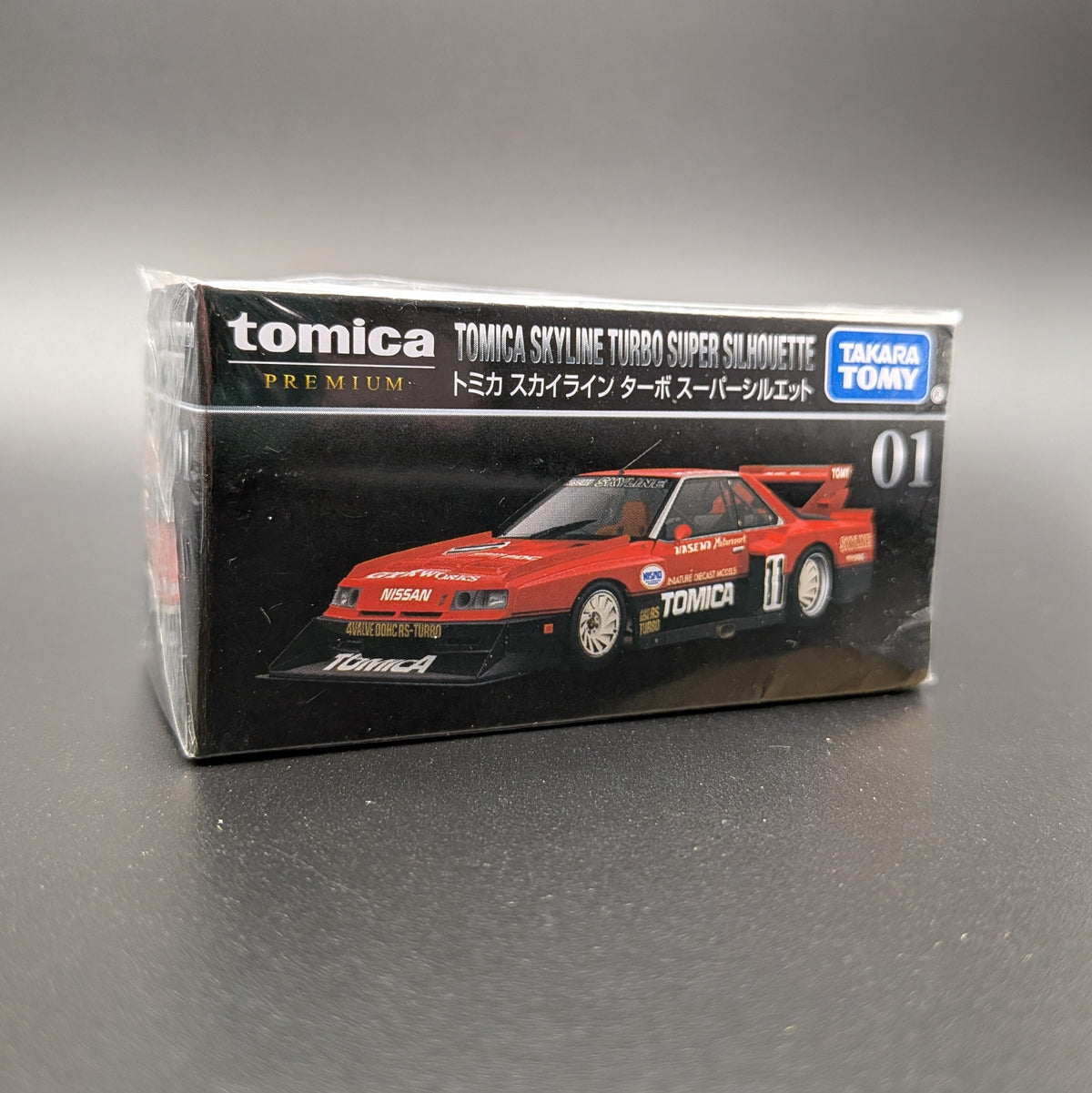 Tomica Premium model car of a Nissan Skyline Turbo Super Silhouette #01 in red and black with Tomica livery. Front facing  at a slight 3/4 angle and on a grey background.