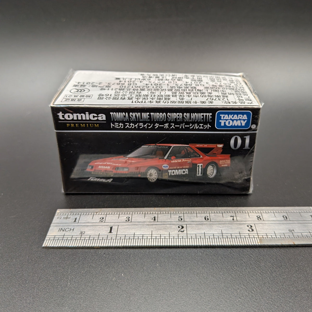 Tomica Premium model car of a Nissan Skyline Turbo Super Silhouette #01 in red and black with Tomica livery. The item is front facing  with a metal ruler beside the box to show the length (8cm) and sits on a grey background.