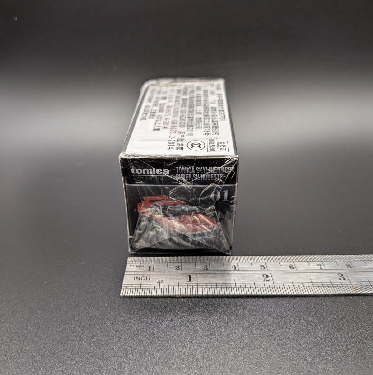 Tomica Premium model car of a Nissan Skyline Turbo Super Silhouette #01 in red and black with Tomica livery. The item is sitting side on  with a metal ruler beside the box to show the width (4cm) and sits on a grey background.