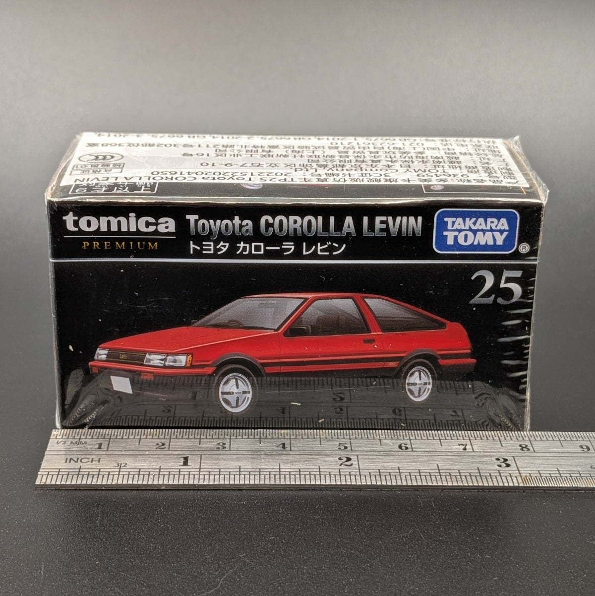 A boxed Tomica Premium toy car model, specifically the Toyota Corolla Levin number 25, shown in its original retail packaging with a metal ruler at the bottom for scale against a dark gray background.