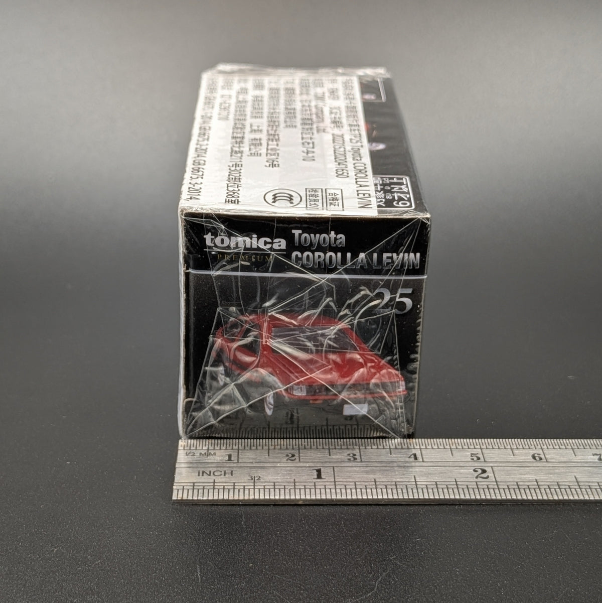 A boxed Tomica Toyota Corolla Levin die-cast model car, still wrapped in clear plastic, shown on a dark gray surface with a metal ruler along the bottom for scale.