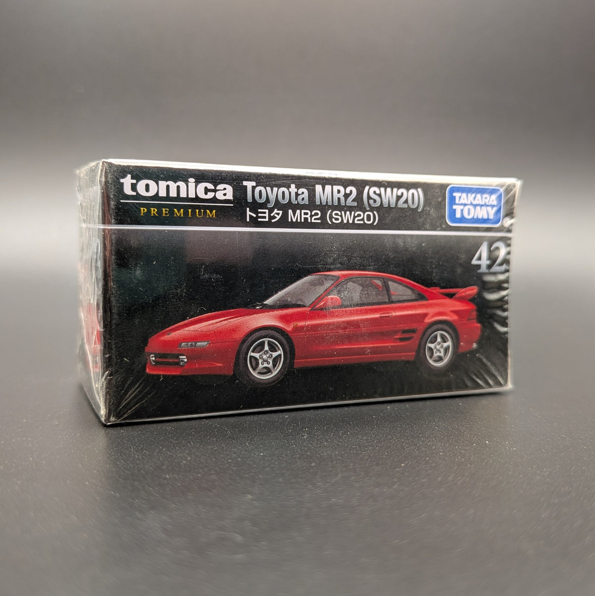 A boxed Tomica Toyota MR2 (SW20) model car package on a dark gray studio background. The front of the box shows a red sports car and visible branding including Tomica, Toyota, and Takara TOMY.
