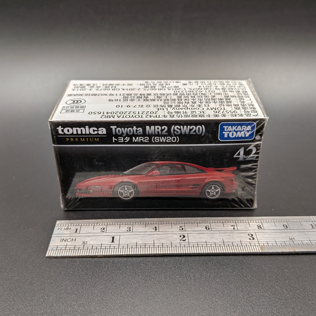 A boxed Tomica Toyota MR2 (SW20) model car on a dark gray background, with a metal ruler placed in front for scale. The retail box shows a red die-cast model car and Takara Tomy branding.