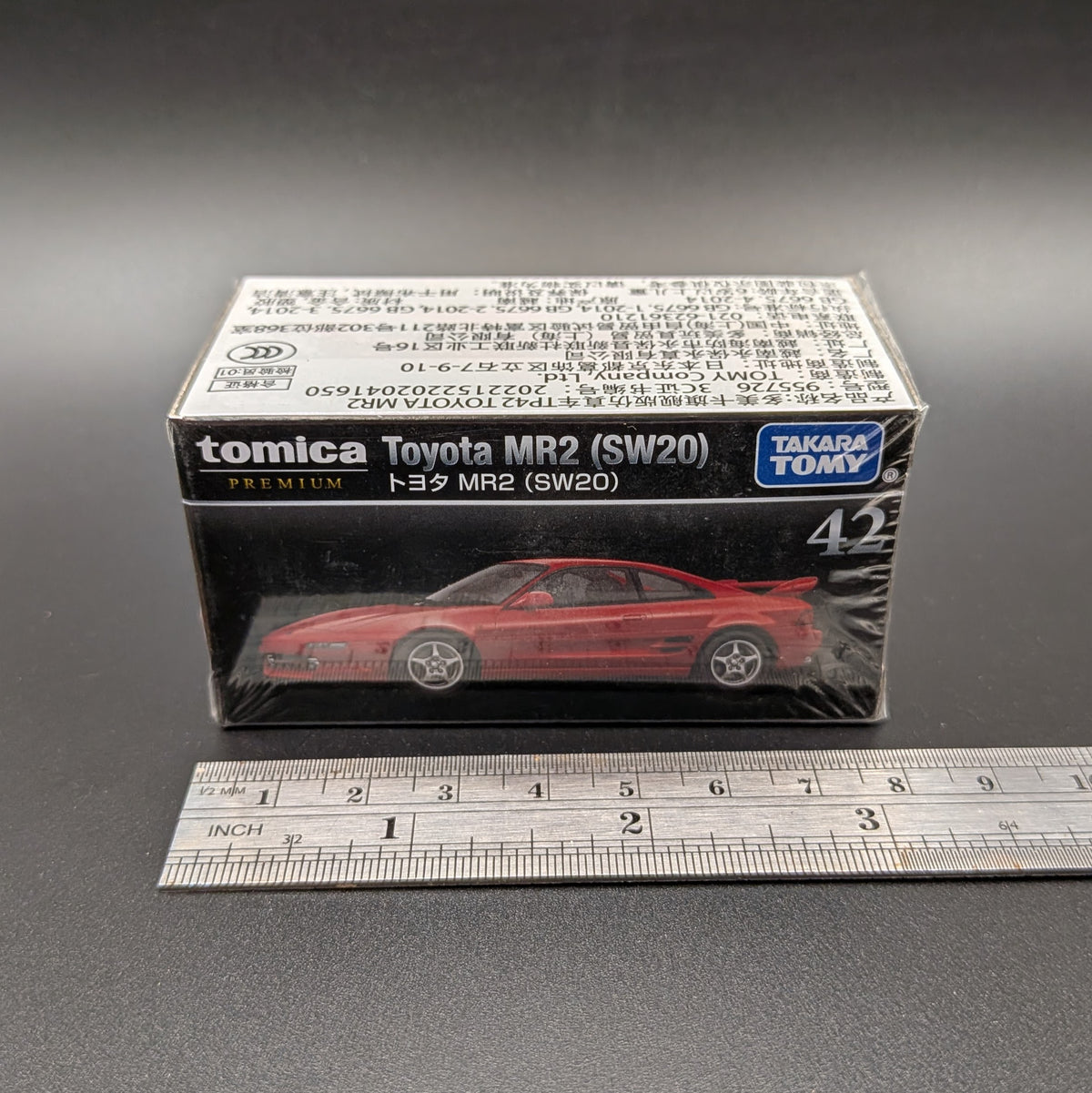 A boxed Tomica Toyota MR2 (SW20) model car on a dark gray background, with a metal ruler placed in front for scale. The retail box shows a red die-cast model car and Takara Tomy branding.
