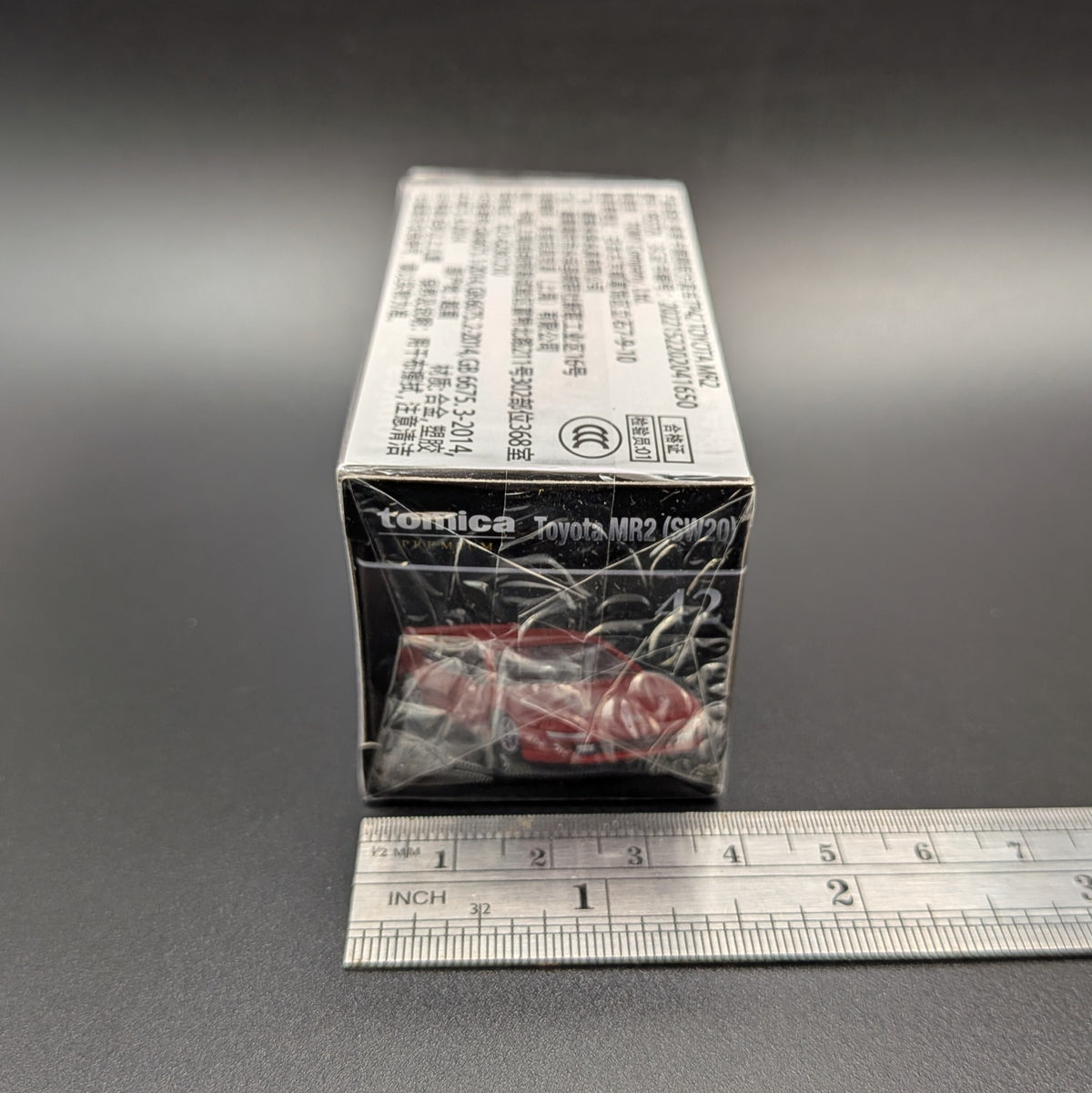 A boxed Tomica Toyota MR2 (SW20) model car package standing on a dark gray surface, with a metal ruler placed in front for scale. The box is mostly black and white with a small red car visible through clear plastic wrapping.