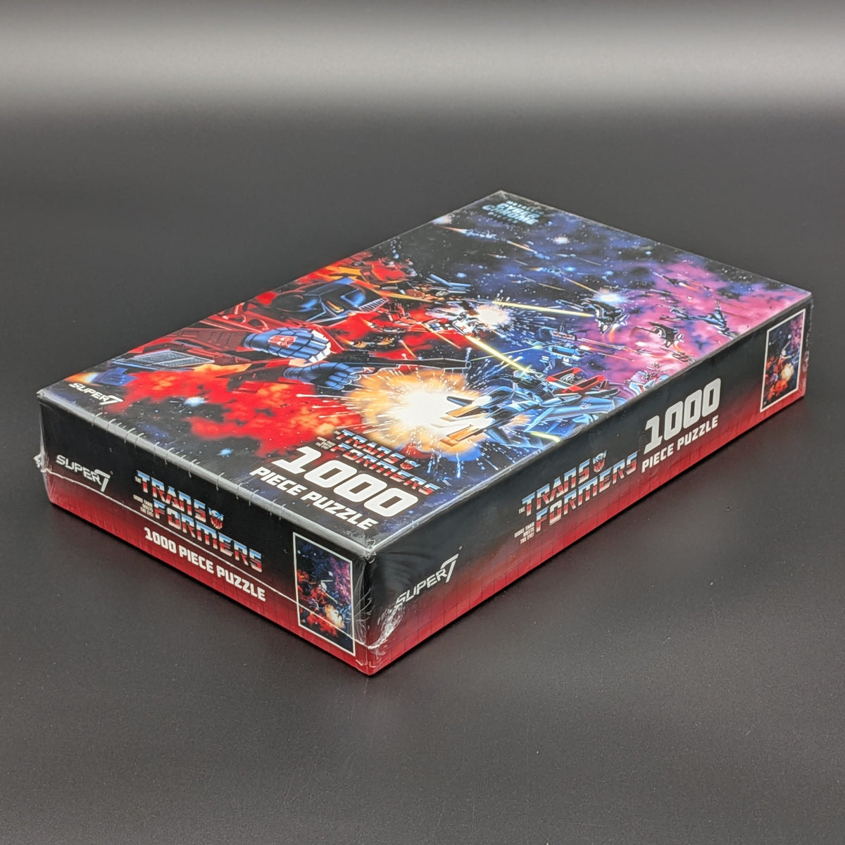 A 1000-piece jigsaw puzzle box by Super7 featuring a colorful space battle scene with Transformers and other robot characters, photographed on a dark gray background.