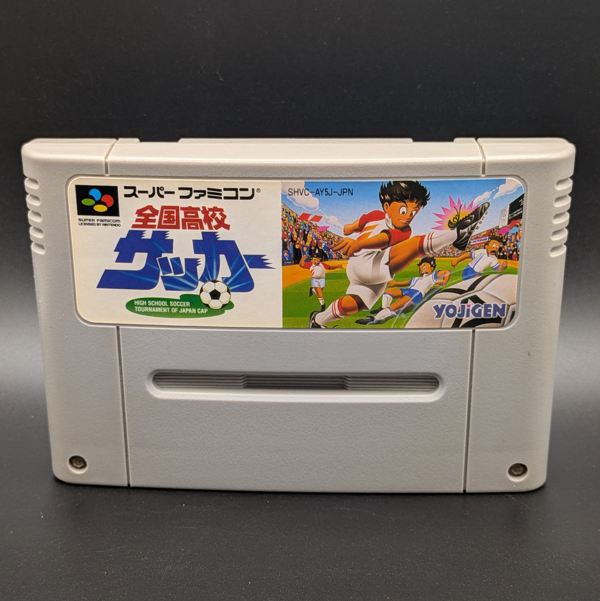 A close up of Zenkoku Kōkō Soccer video game cartridge for Nintendo's Super Famicom (SFC) Entertainment System, shown standing up and facing forwards on a dark grey background.