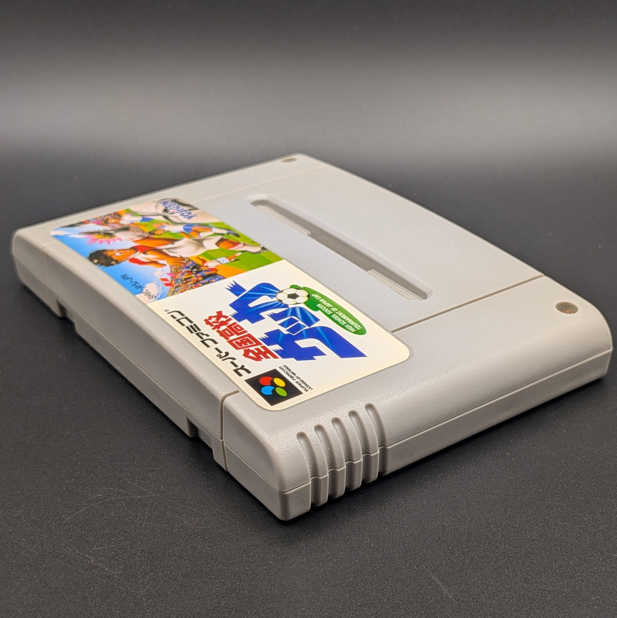 Zenkoku Kōkō Soccer video game cartridge for Nintendo's Super Famicom (SFC) Entertainment System, shown laying on its back with the label facing upwards. Showing the left hand side and top edge. It sits on a dark grey background.