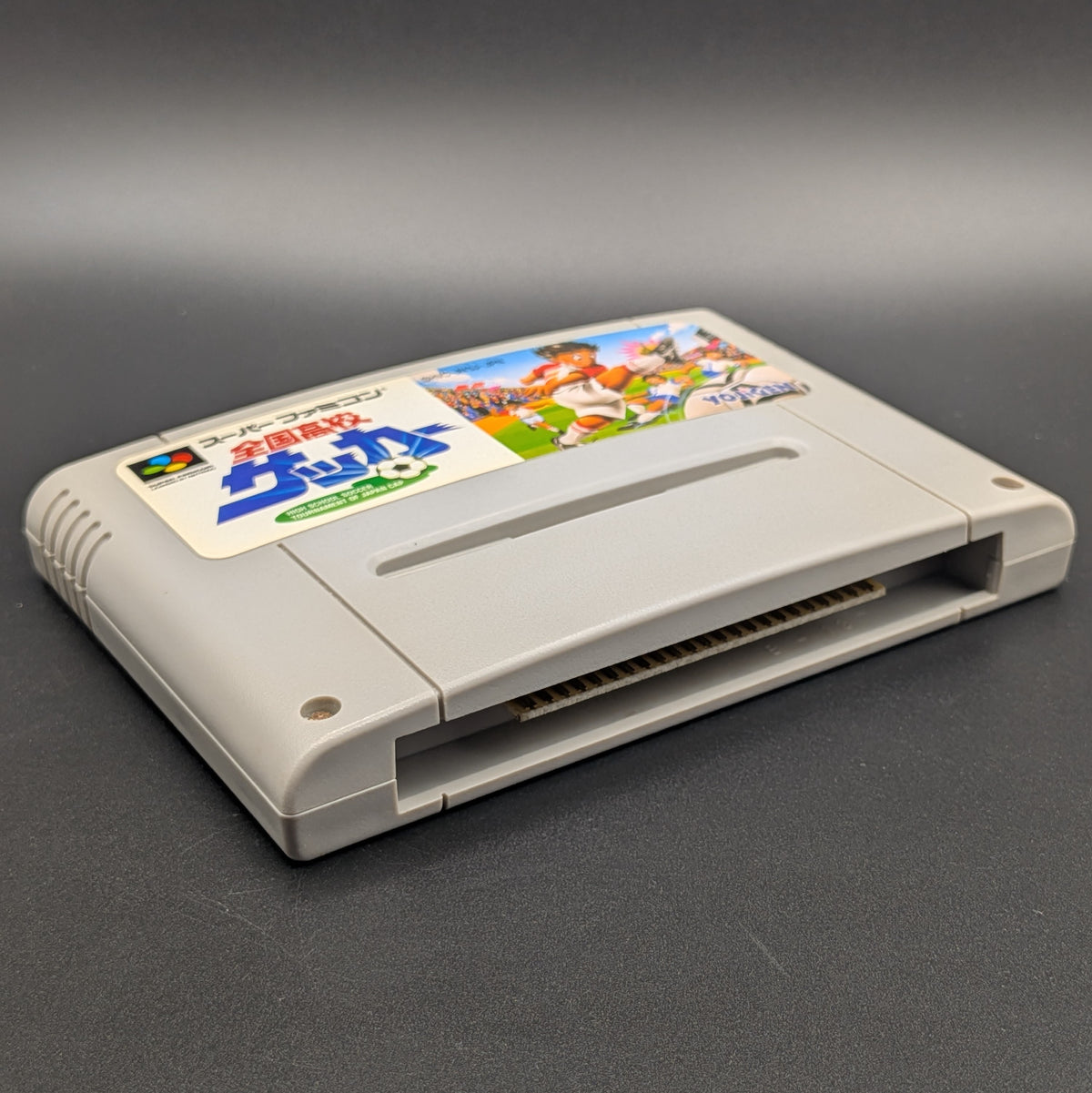Zenkoku Kōkō Soccer video game cartridge for Nintendo's Super Famicom (SFC) Entertainment System, shown laying on its back with the label facing upwards. Showing the left hand side and bottom slot. It sits on a dark grey background.
