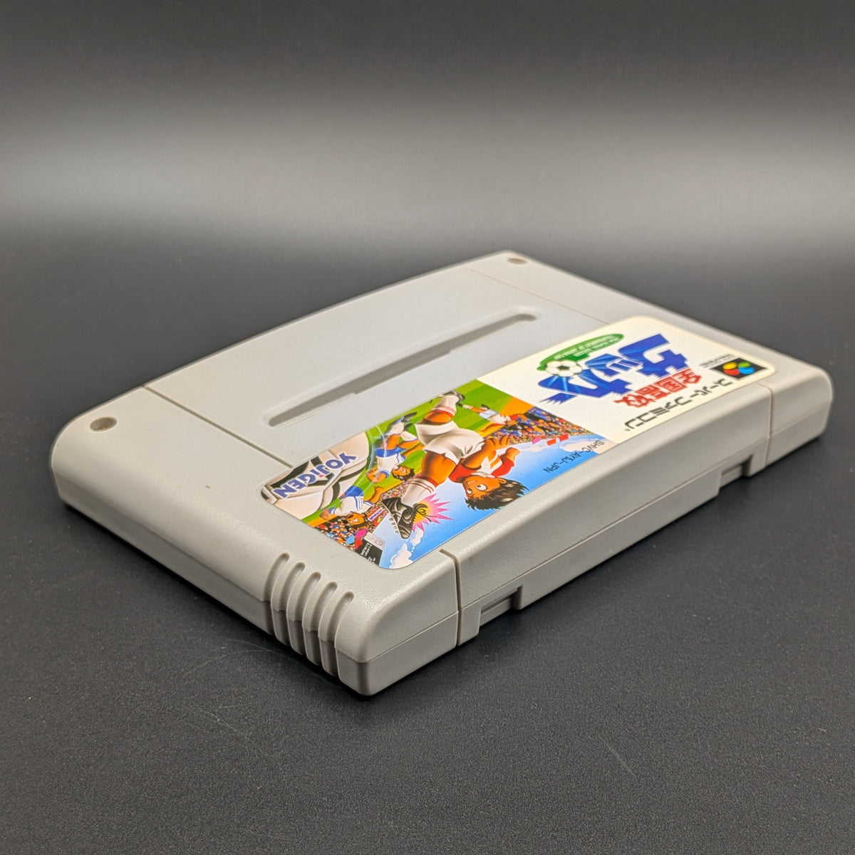 Zenkoku Kōkō Soccer video game cartridge for Nintendo's Super Famicom (SFC) Entertainment System, shown laying on its back with the label facing upwards. Showing the right hand side and top edge. It sits on a dark grey background.