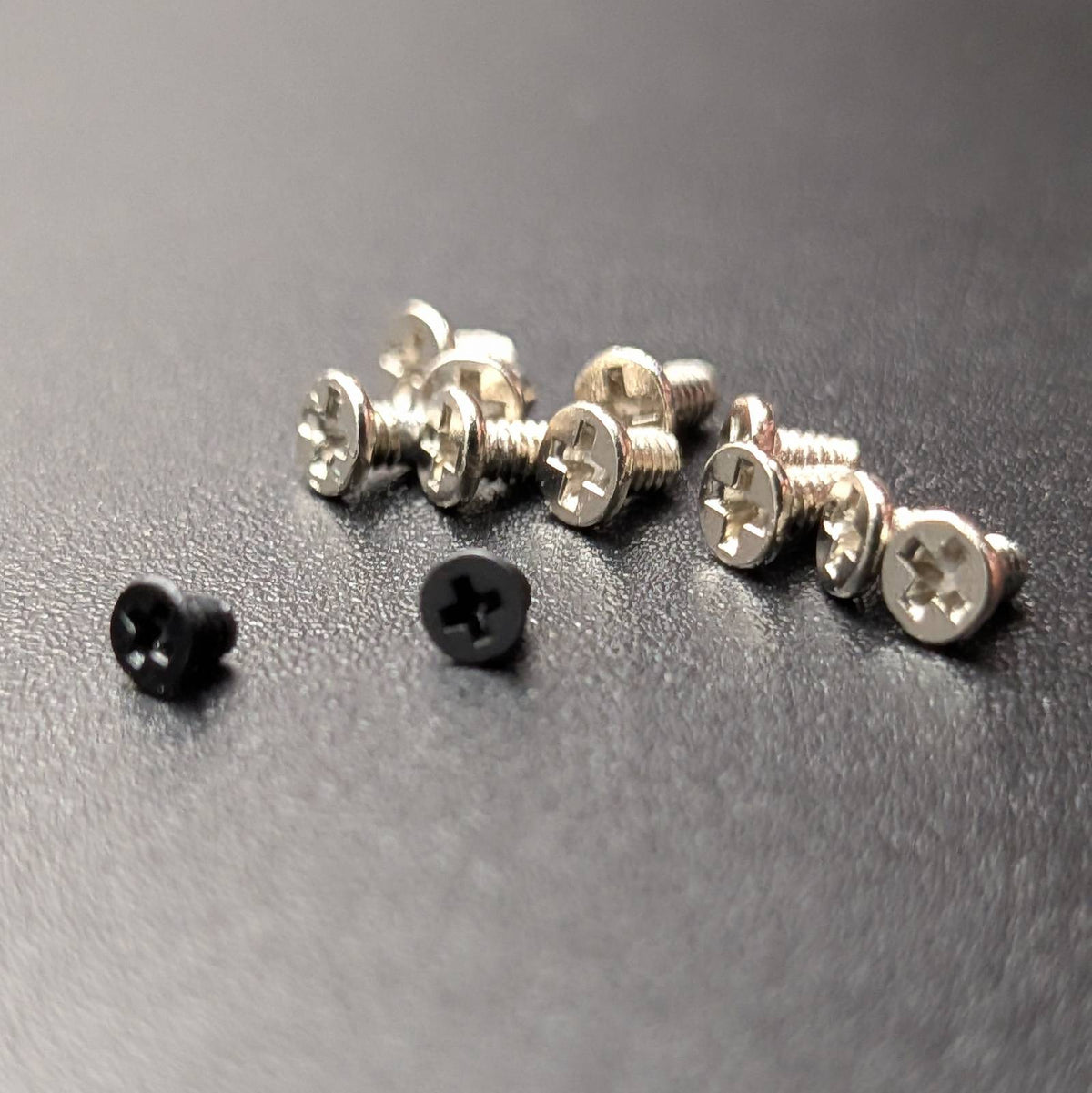 Twelve small screws on a dark gray textured surface, including ten silver screws and two black screws.