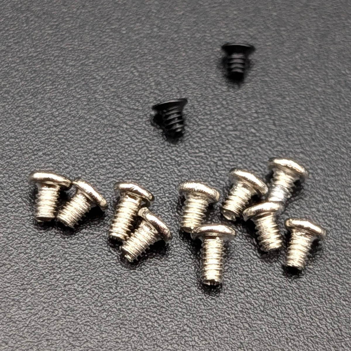 Twelve small metal screws with flat round heads and threaded shanks arranged on a dark textured surface.