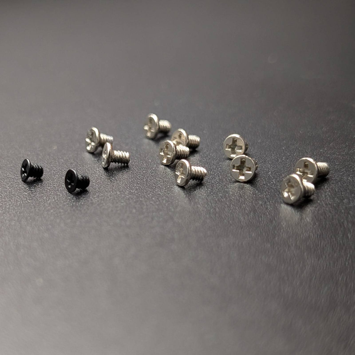 A set of small screws, including two black screws and several silver screws, arranged on a dark gray textured surface.