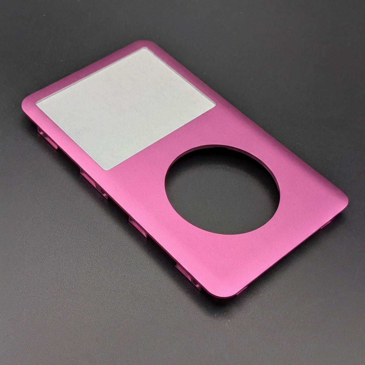 A metallic pink shell designed for an iPod Classic, featuring a cutout for the circular touchpad and side button controls, shown on a dark gray background.