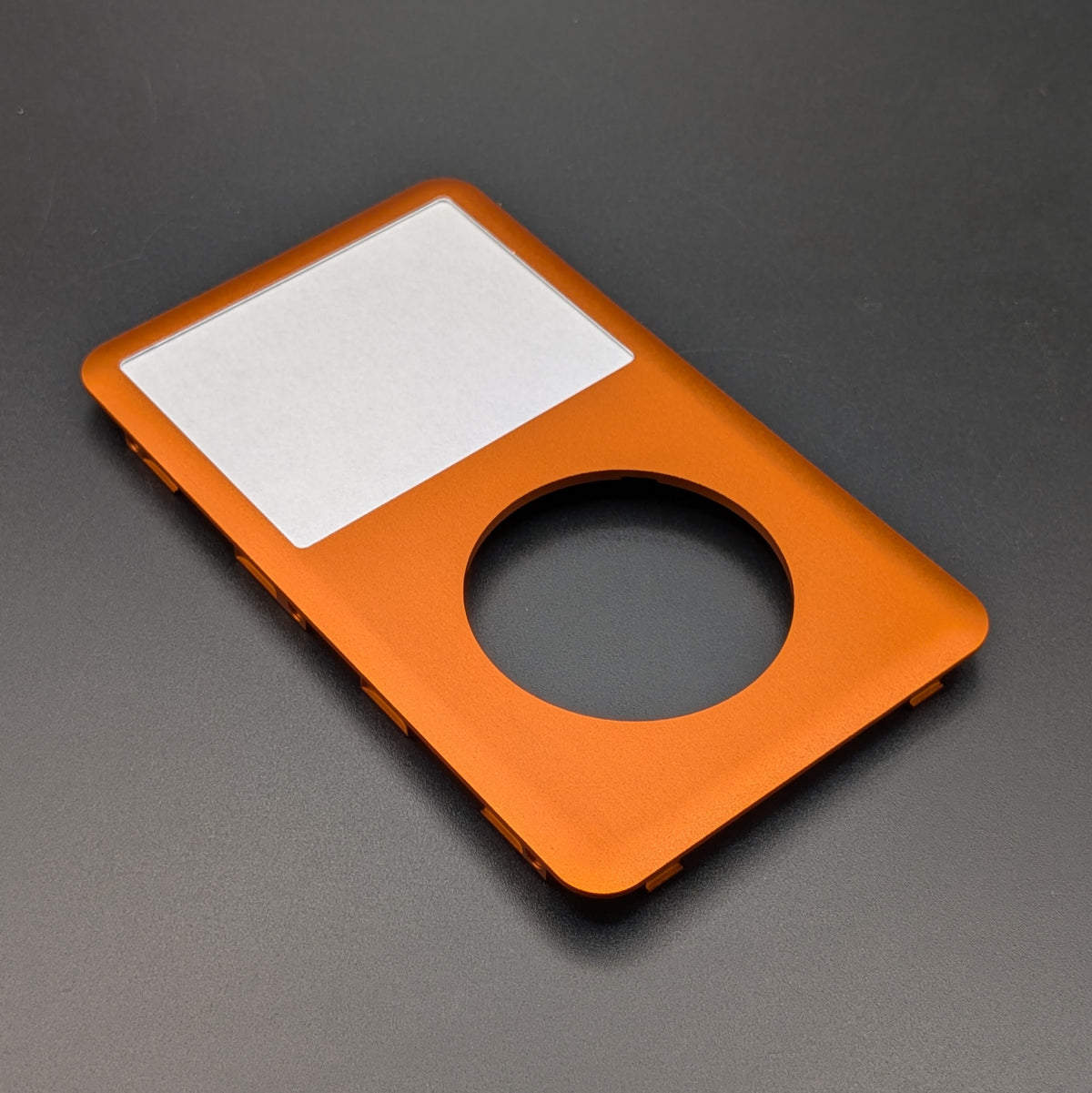 An orange aluminum replacement bezel or faceplate for an iPod Classic, featuring a cutout for the screen and a circular opening, shown on a dark gray background.