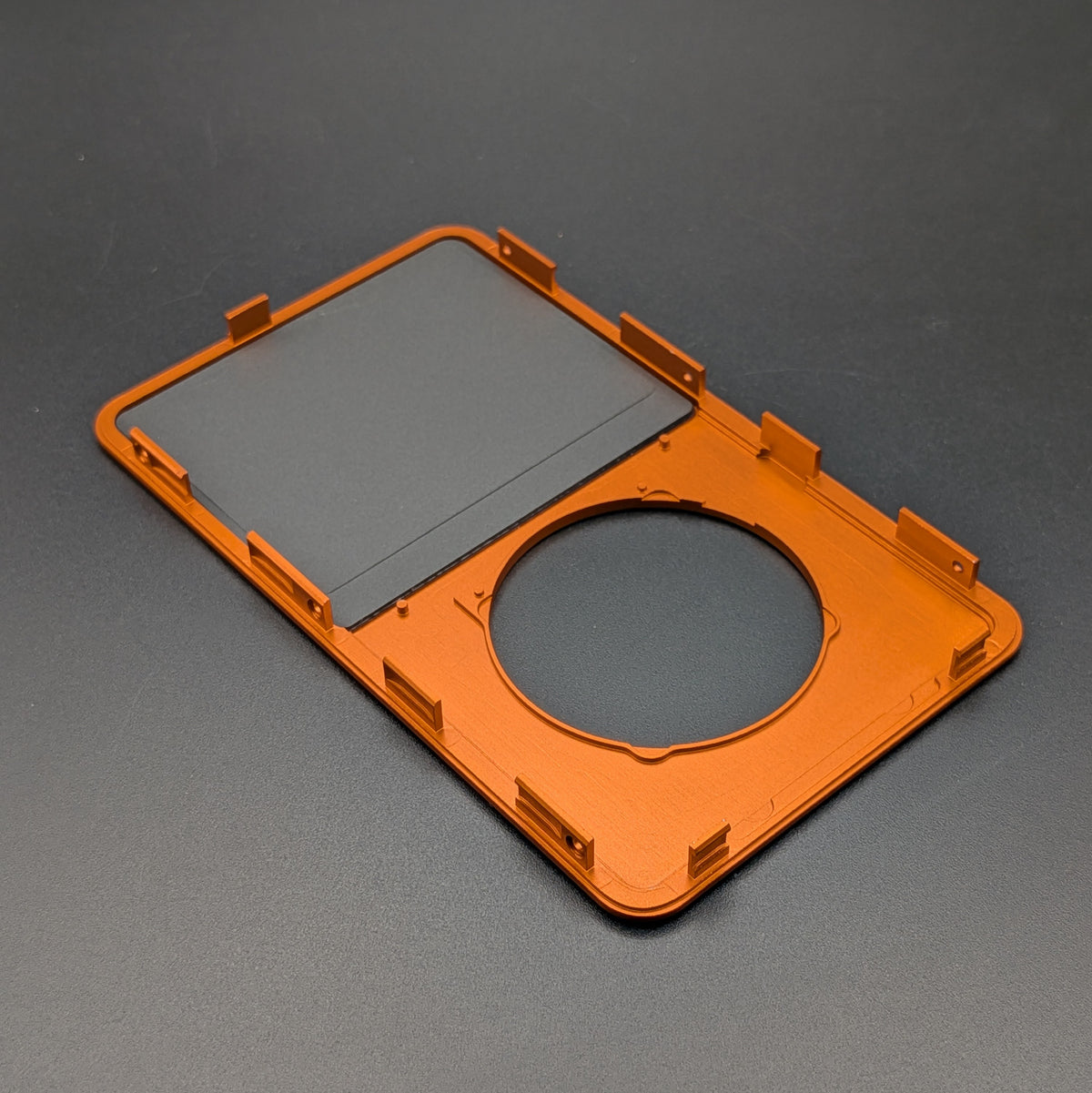 An orange protective frame or cover with a circular cutout and a rectangular dark insert, shown on a dark gray textured background.