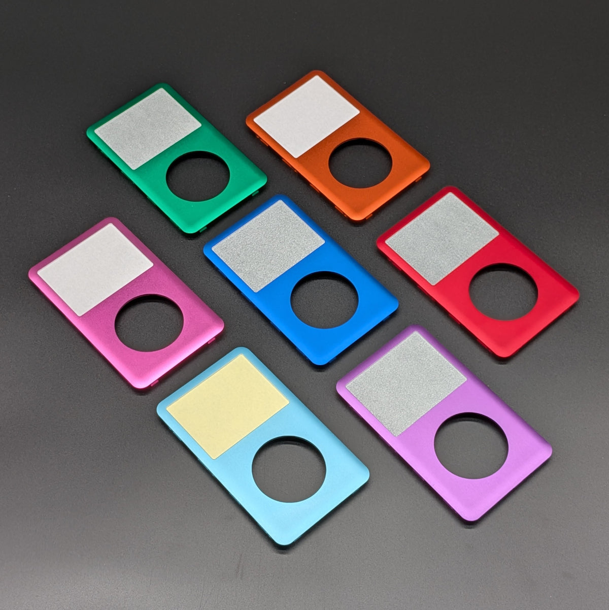 Six rectangular metal accessory pieces in different colors are arranged on a dark gray background. Each piece has a circular cutout and a square textured panel, shown in green, orange, red, pink, blue, light blue, and purple.