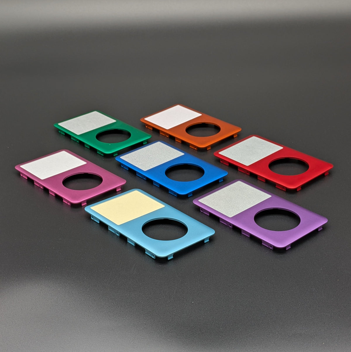 Six colorful rectangular metal components with circular cutouts and silver screens, shown in green, orange, red, pink, blue, and purple on a dark gray background.