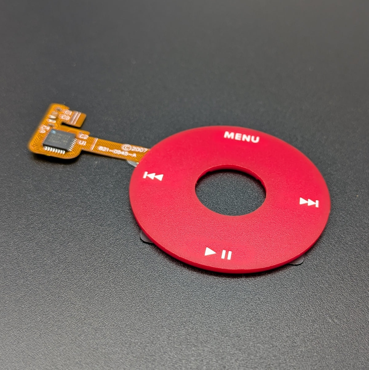 A red circular electronic control module with white playback symbols and the word 'MENU' on it, attached to a small orange flex cable, photographed on a dark gray textured surface.