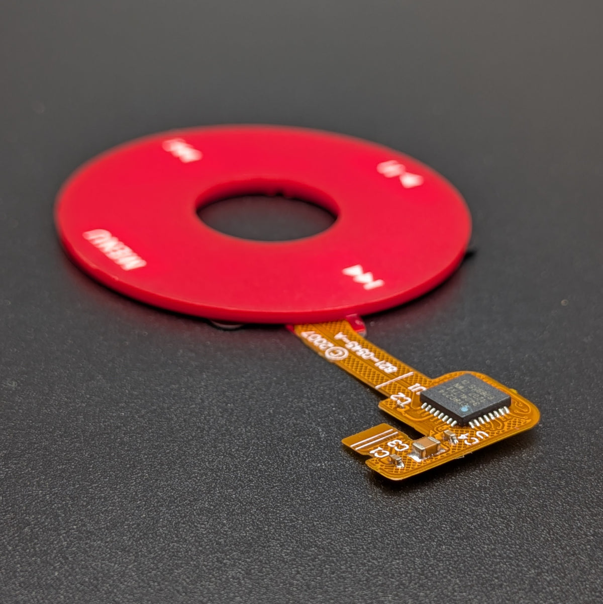 A red circular ring-shaped electronic component with a flexible flex cable leading to a small integrated circuit chip, photographed on a dark textured surface.