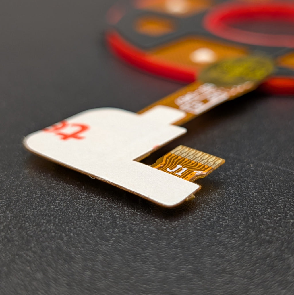 A close-up of a replacement flex circuit component with a white protective cover and gold-plated contacts, resting on a dark textured surface.