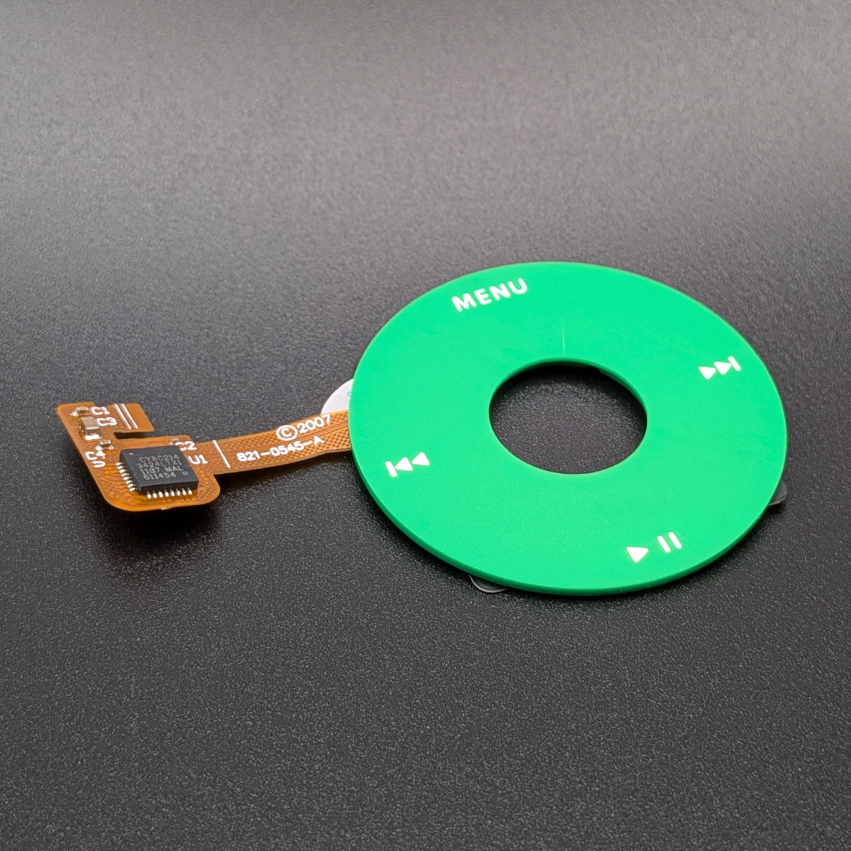 A green circular media control button pad with white printed icons and the word 'MENU', attached to a small orange flex cable with a black chip, photographed on a dark gray surface.