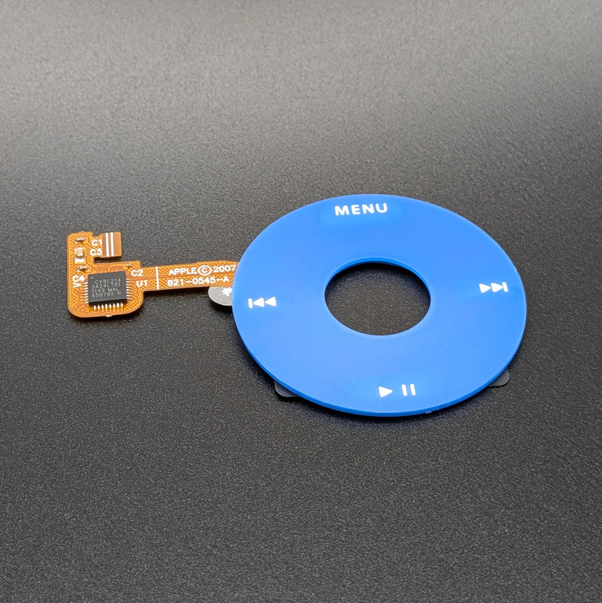 A blue circular Apple iPod media button with white control symbols and the word MENU, shown next to a small orange flex cable on a dark gray textured background.