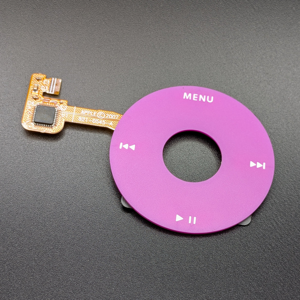 A replacement control panel for an Apple iPod, featuring a circular magenta purple faceplate with white playback controls and the word 'MENU' printed on it, connected to a gold flex cable with a black microchip, set against a dark gray textured background.