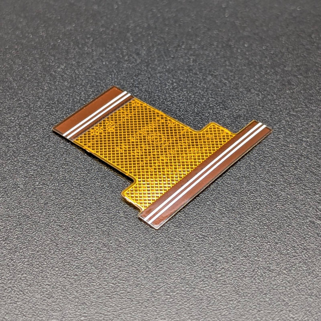 A small flexible circuitry connector piece with a gold mesh center and brown copper traces, photographed on a dark textured surface.
